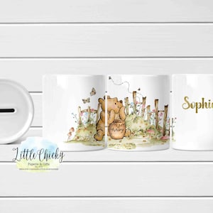 Puede incluir: Juego de hucha y taza de cerámica blanca. La taza presenta una ilustración de Winnie the Pooh con un tarro de miel. La hucha tiene una ranura para insertar monedas. La taza está personalizada con el nombre "Sophia" en dorado.