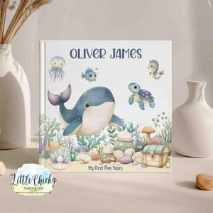 May include: A personalized baby book with the name "Oliver James" in navy blue script. The cover features watercolor illustrations of a whale, turtle, seahorse, and other sea creatures. The text "My First Five Years" is also included.