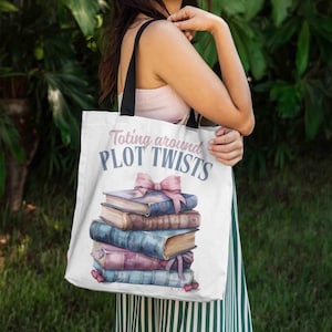May include: White tote bag with black straps. The bag has the text "Toting around PLOT TWISTS" in blue and pink. It features a stack of books with pink bows. The books are in shades of blue, brown, and pink.