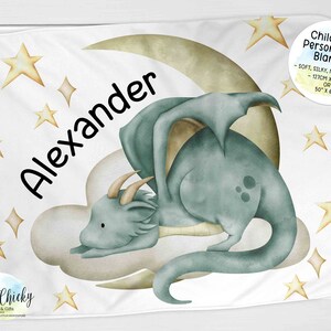 May include: A white personalised blanket featuring a blue dragon sleeping on a crescent moon. The blanket has a yellow star pattern and the name "Alexander" is written in black. The blanket is 50 inches by 60 inches.