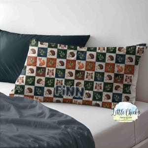 May include: A rectangular pillowcase with a woodland animal pattern. The design features a checkerboard pattern with foxes, owls, and hedgehogs. The name "Finn" is printed in blue letters. The pillowcase is on a white bed.