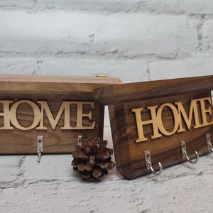 Könnte beinhalten: Zwei Schlüsselhalter aus Holz mit der Gravur "HOME". Jeder Halter hat drei Haken zum Aufhängen von Schlüsseln. Die Halter sind aus dunkelbraunem Holz und haben einen rustikalen Look.
