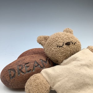May include: A brown teddy bear wearing a beige shirt is lying down next to a wooden heart with the word "DREAM" carved into it.