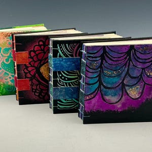 May include: Four square journals with exposed stitch bindings. The covers feature vibrant, hand-painted designs in green, orange, red, blue, and purple, with black accents, intricate patterns, and visible gold-toned glitter details.