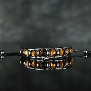 Puede incluir: Una pulsera con cuentas de ojo de tigre y detalles metálicos plateados. Las cuentas son de color marrón cálido y la pulsera tiene un diseño tejido. La pulsera se muestra sobre una superficie reflectante.