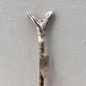 May include: A walking stick featuring a Y-shaped antler handle with a white and brown textured surface. The wooden shaft has a mottled brown pattern and is attached to a brown paper tag with the handwritten number 18 and a small bird illustration.