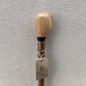 May include: A wooden walking stick featuring a light-coloured, smooth, rounded handle attached to a darker, textured shaft. A black band separates the handle from the shaft. A brown paper tag with the handwritten number 6 is attached by a white string.