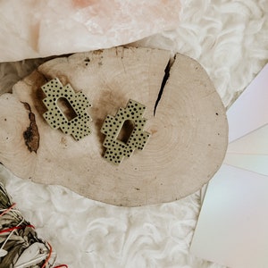 Op de afbeelding: Een paar olijfgroene oorbellen met zwarte stippen, liggend op een lichte houten schijf. De oorbellen hebben een geometrisch ontwerp met een open boog. Een saliebundel en een kleurenwaaier zijn ook zichtbaar.