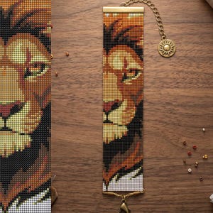 May include: A rectangular beaded bracelet featuring a detailed pixelated lion face pattern in shades of orange, brown, yellow, black, and white. The bracelet has gold-toned metal end bars, a gold chain extension, and a sun-shaped charm.