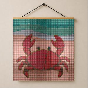 May include: A square diamond painting wall hanging featuring a red crab on a light pink sandy beach with a turquoise ocean and white foam waves. The artwork is composed of small, uniform circular beads and is suspended by a wooden frame hanger.