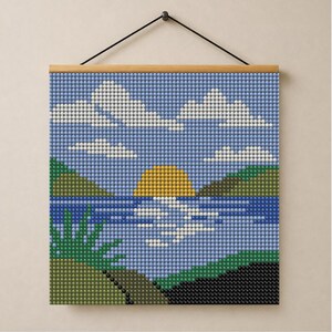 Op de afbeelding: Een vierkante diamond painting wandhanger met een landschap van een gele zon die ondergaat boven een blauw meer, groene heuvels en witte wolken. Het kunstwerk bestaat uit kleine, ronde, gefacetteerde kralen in een raster met een houten lijst.