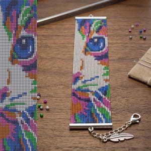 Puede incluir: Una pulsera rectangular de cuentas con un diseño de cara de gato pixelado en colores vibrantes y un ojo azul prominente. La pulsera está terminada con terminales de metal plateado, una cadena extensora y un pequeño dije de pluma plateada.