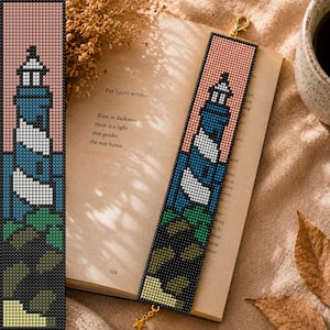 May include: A rectangular beaded bookmark featuring a lighthouse design with a blue and white tower against a pink sky. The base shows green foliage and dark rocks. A gold-toned metal lobster clasp and a small star charm are attached to the top.