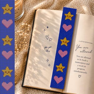 Op de afbeelding: Een rechthoekige boekenlegger met een herhalend patroon van gele sterren met lachende gezichten en lichtroze harten op een donkerblauwe achtergrond, geplaatst in een open boek met de tekst A LITTLE REMINDER You are allowed.