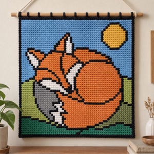 May include: A square crochet wall hanging featuring a pixelated design of a curled-up orange fox with white accents on a green grassy hill under a light blue sky with a yellow sun. The piece is framed by a black border and hangs from a wooden rod.