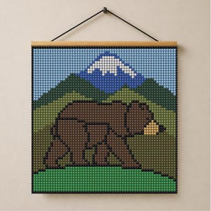 May include: A square wall hanging featuring a pixelated bead art design of a brown bear walking across a green field. In the background, there are green hills and a blue mountain with a white snow-capped peak. The art is held by a wooden frame.