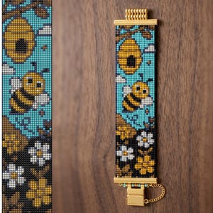 May include: A rectangular beaded bracelet featuring a pattern of a yellow beehive, a striped bee, white flowers, and brown leaves against a bright turquoise and black background. The bracelet has gold-toned metal end bars and a matching clasp.