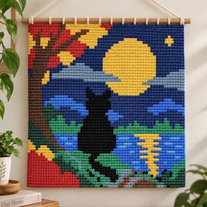 Puede incluir: Un tapiz de ganchillo cuadrado que muestra la silueta de un gato negro frente a una gran luna llena amarilla, un cielo nocturno azul oscuro, un árbol con hojas rojas y amarillas, colinas verdes y un lago azul que refleja la luz lunar.