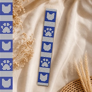 May include: A blue and white beaded bookmark with a repeating pattern of cat faces and paw prints. The bookmark is rectangular and has a vertical design. The background is a neutral beige fabric.