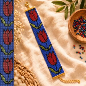 Op de afbeelding: Een kralenarmband met een herhalend patroon van rode tulpen met groene stelen en bladeren op een blauwe achtergrond. De armband is afgewerkt met goudkleurige metalen eindstukken, een karabijnsluiting en een verstelbare kettingverlenging.