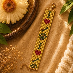 May include: A rectangular beaded bracelet featuring a yellow background with a repeating pattern of the letter I, a red heart, and a dark green paw print. The bracelet is finished with gold-toned metal hardware, including a lobster claw clasp.