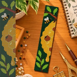 May include: A rectangular beaded bookmark featuring a dark green background with a yellow honeycomb pattern, green leaves on brown branches, and a small bee. The design is created with small, uniform beads arranged in a grid-like pattern.