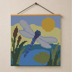 May include: A square diamond painting wall hanging featuring a blue dragonfly hovering over a pond with green lily pads and cattails. The scene includes a yellow sun in a light blue sky with white clouds, all rendered in a grid of small round beads.