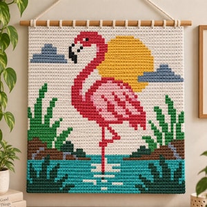 Puede incluir: Un tapiz de ganchillo rectangular que muestra un flamenco pixelado en tonos rosa y rojo de pie en agua azul. La escena incluye un sol amarillo, nubes grises y follaje verde sobre un fondo crema, suspendido de una varilla de madera.