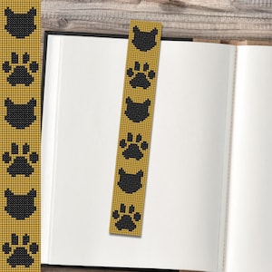 May include: A gold-coloured beaded bookmark featuring a repeating pattern of black cat silhouettes and paw prints. The bookmark is in an open book with white pages, on a wooden surface.