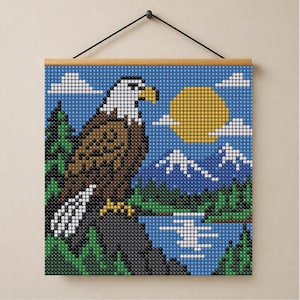 May include: A square diamond painting wall hanging featuring a pixelated bald eagle perched on a rock. The scene includes a yellow sun, white clouds, blue sky, green pine trees, and a blue lake with snow-capped mountains in the background.