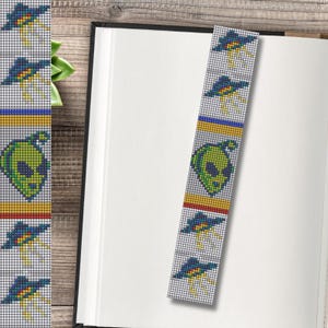 May include: A handmade bookmark featuring a repeating pattern of a green alien head and blue UFOs. The bookmark has a white background with yellow, blue, and red stripes. The bookmark is placed in an open book.