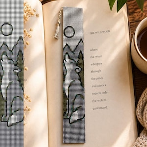 May include: A rectangular beaded bookmark featuring a pixelated design of a wolf howling at a circular moon against a backdrop of mountains and pine trees. The bookmark is crafted from small grey, white, and green beads and includes a small silver tassel.