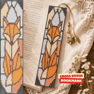 May include: A rectangular cross-stitch bookmark featuring a fox design in orange, cream, and black, styled like stained glass. It includes a gold-toned cord with a small metal fox charm and a leaf-shaped pendant attached to the top eyelet.