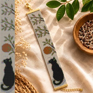 Könnte beinhalten: Ein rechteckiges Perlenarmband mit einem sich wiederholenden Muster aus einer schwarzen Katze, einer runden orangefarbenen Frucht und grünen Blattzweigen auf weißem Grund. Das Armband hat goldfarbene Metallendkappen, einen Karabinerverschluss und eine Verlängerungskette.