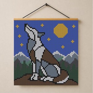 May include: A square wall hanging featuring a pixelated mosaic design of a wolf howling at a yellow moon. The scene includes a blue starry night sky, snow-capped mountains, and a dark forest landscape, all suspended from a wooden rod.