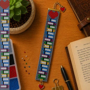 May include: A rectangular beaded bookmark featuring a gray background with a pattern of colorful horizontal lines and a red heart at the top. The bookmark includes a gold-toned metal chain attachment with a small red heart charm at the end.