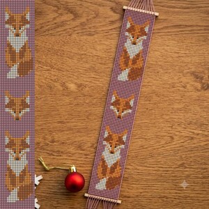 Könnte beinhalten: Ein langer, rechteckiger Wandbehang aus Perlen mit einem sich wiederholenden Muster aus orangefarbenen, weißen und braunen Füchsen auf violettem Grund. Das Stück hängt an einem Holzstab und hat am unteren Rand eine Fransenborte.