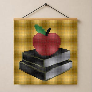 May include: A square wall hanging featuring a pixelated design of a red apple with a green leaf resting on two stacked books. The books have black covers and light gray pages. The background is a solid yellow grid pattern with a wooden frame.