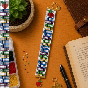 May include: A rectangular beaded bookmark featuring a white background with a vertical pattern of colorful stacked books in blue, green, yellow, and purple. A red apple charm hangs from the bottom, and a heart-shaped gold clasp is at the top.