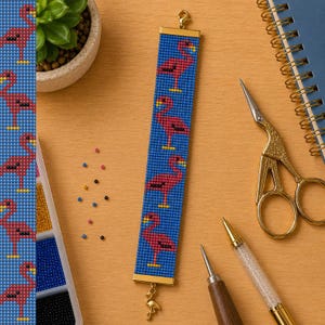 May include: A rectangular beaded bracelet featuring a repeating pattern of pink flamingos on a blue background. The bracelet has gold-toned metal end bars and a small gold flamingo charm dangling from the bottom. It is crafted with small beads.