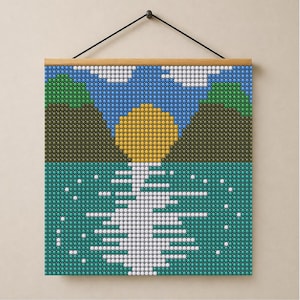 Op de afbeelding: Een vierkante wandhanger met een gepixeld landschapsontwerp met een gele zon, blauwe lucht, witte wolken, groene bergen en een blauwgroen wateroppervlak met een witte reflectie van de zon, opgehangen aan een houten lat en een zwart koord.
