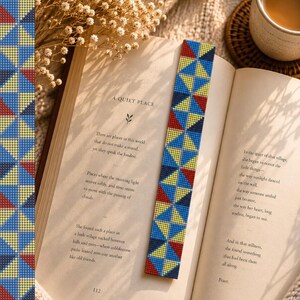 May include: A patterned bookmark featuring a geometric design in blue, yellow, and red, inserted into an open book. The book's title, "A Quiet Place", is visible. The background includes a cup of tea and dried flowers, creating a cozy reading scene.