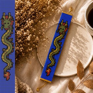 May include: A rectangular beaded bracelet featuring a pixelated green dragon design on a vibrant blue background. The bracelet is finished with gold-toned metal end caps, a lobster claw clasp, and a small star-shaped charm attached to the chain.
