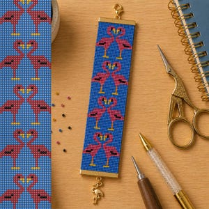 Op de afbeelding: Een rechthoekige kralenarmband met een herhalend patroon van roze flamingo's op een blauwe achtergrond. De armband heeft goudkleurige metalen eindstukken en een kleine gouden flamingo-bedel aan de onderkant. Het kralenwerk is uniform.
