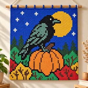 Op de afbeelding: Een vierkante gehaakte wandhanger met een zwarte kraai op een oranje pompoen tegen een donkerblauwe nachtelijke hemel met een gele volle maan en witte sterren. De scène bevat groene en rode bomen aan de basis en hangt aan een houten roede.