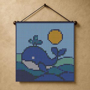 May include: A square wall hanging featuring a pixelated mosaic design of a dark blue whale swimming in light blue and teal water under a bright yellow sun. The artwork is constructed from small circular beads and suspended by a black cord.