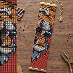 May include: A rectangular beaded bracelet featuring a detailed tiger face pattern in orange, black, white, and grey beads. The bracelet is finished with gold-toned metal end bars and a gold-toned chain featuring a small tiger head charm at the end.