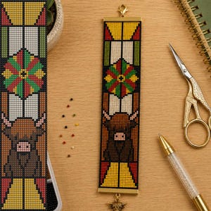 May include: A rectangular beaded bookmark featuring a stained glass pattern with a brown highland cow at the bottom, a geometric floral motif in red, green, and yellow in the centre, and a gold metal clasp at the top with a small cow charm.