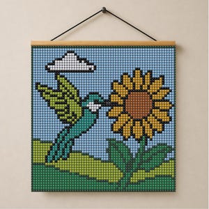 Puede incluir: Un tapiz cuadrado con un diseño de arte en cuentas pixeladas de un colibrí verde cerca de un girasol amarillo. La escena incluye una nube blanca sobre un cielo azul claro y una colina verde, suspendido por una varilla de madera.