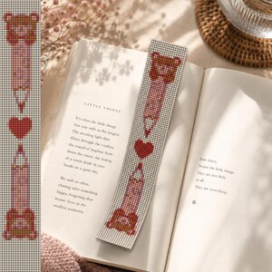 Puede incluir: Un marcapáginas de cuentas con un diseño de lápiz y corazón. El marcapáginas es principalmente blanco con detalles rojos, marrones y rosas. Se encuentra en un libro abierto con el texto "LITTLE THINGS" visible.